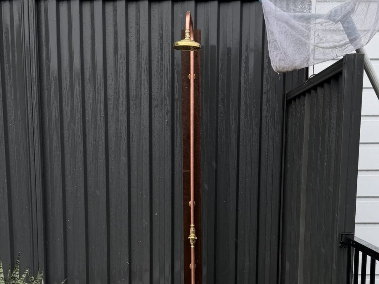 Outdoor shower with an exposed copper riser and brass rose beside a rendered planter