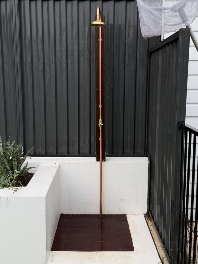 Outdoor shower with an exposed copper riser and brass rose beside a rendered planter